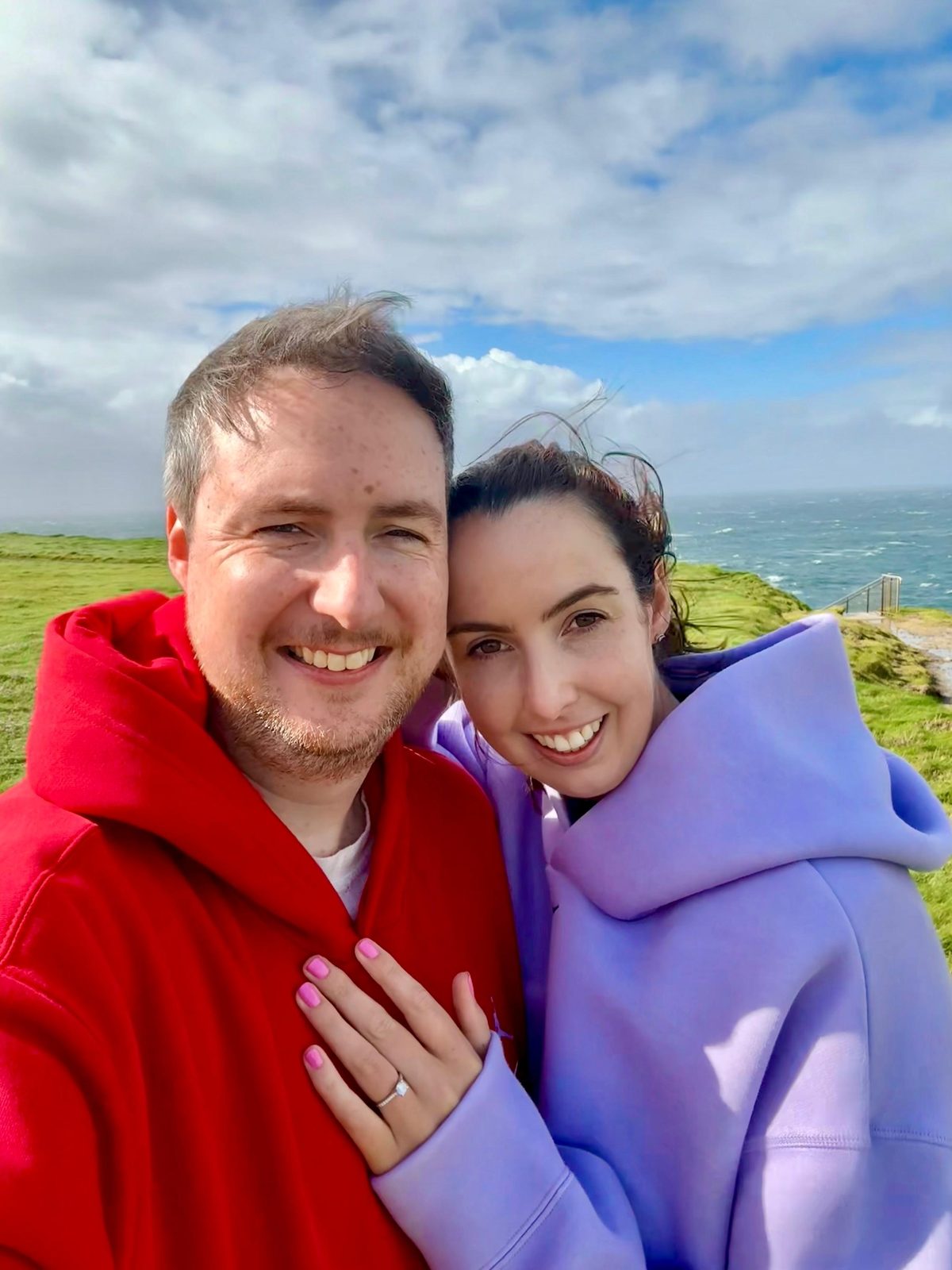 A selfie of Niall in a red hoodie and Orlaith in a lilac hoodie in Kilkee, minutes after the proposal, her ring on show and the Atlantic behind